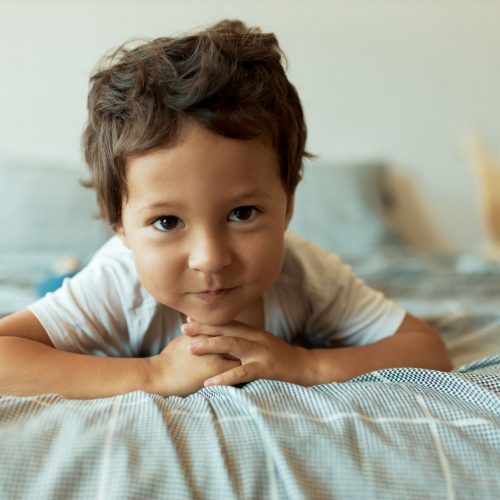 healthy-charming-3-year-old-latin-toddler-lying-rumpled-sheets-with-hands-clasped-front-him-having-curious-play-fun-facial-expression