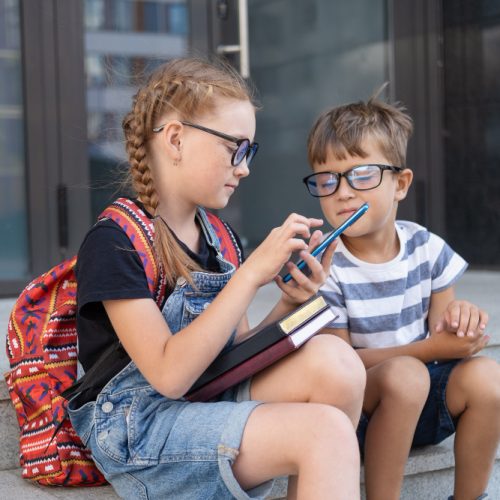 two-cute-caucasian-happy-kids-eyeglasses-with-books-backpack-sit-talking-typing-phone-after-school-knowledge-day-back-school-concept